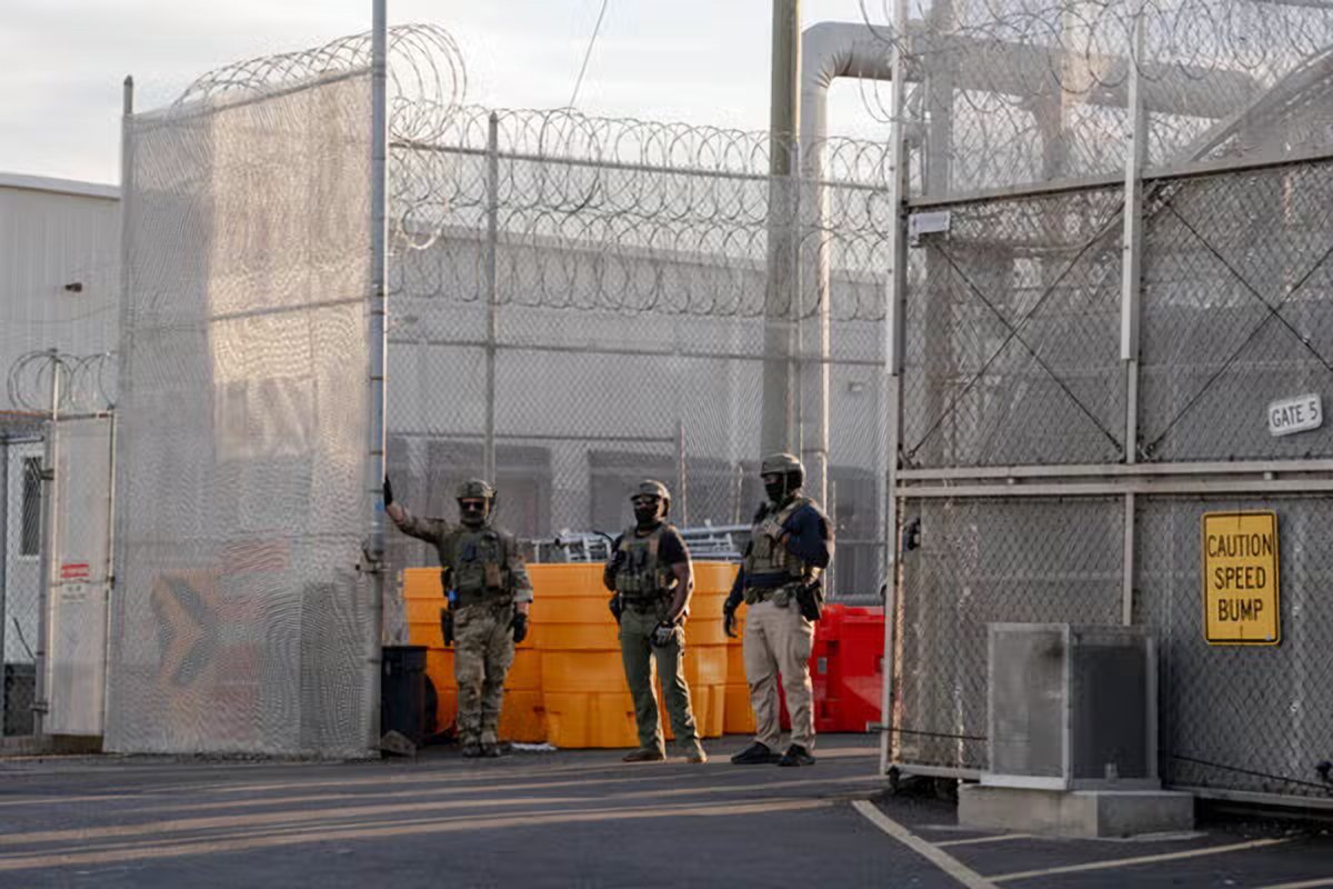 Three federal agents in tactical gear stand inside a fenced gate topped with razor wire at Delaney Hall.