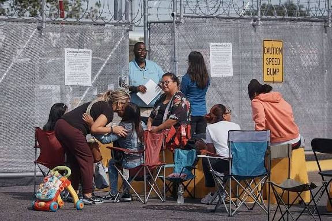 Families sit in folding chairs outside the fence at Delaney Hall, under a posted contraband notice.