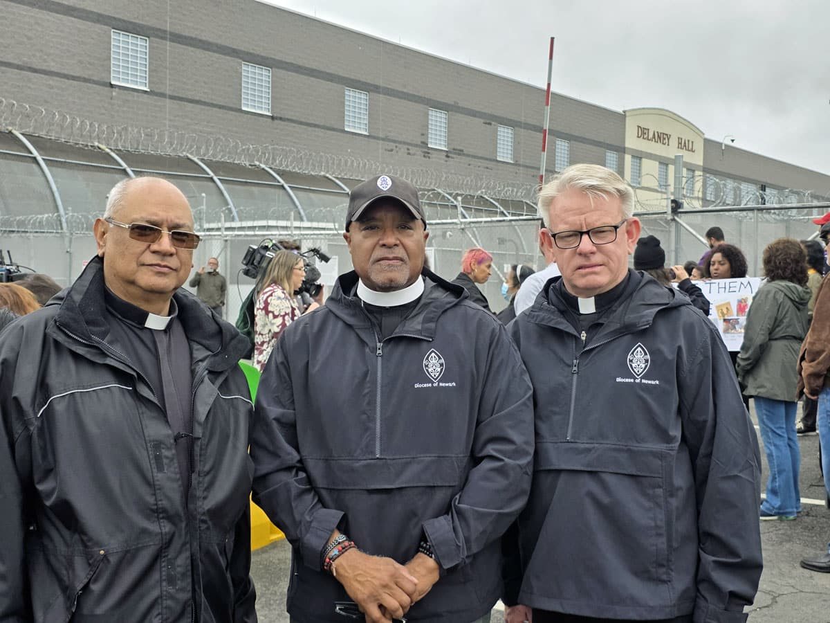 Three clergy members in Diocese of Newark jackets stand in front of the Delaney Hall fence and building.