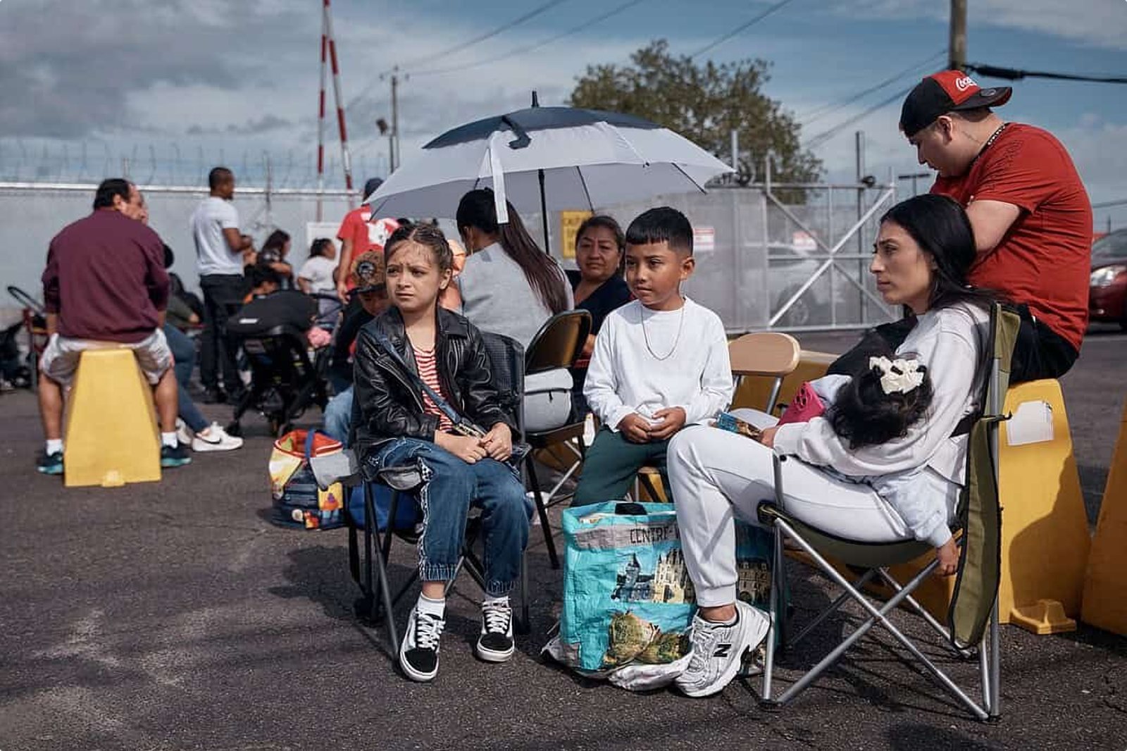 Families sit in folding chairs in the parking lot outside the fence at Delaney Hall. A girl in a leather jacket sits beside a boy in white, and a mother holds a sleeping toddler under an umbrella.