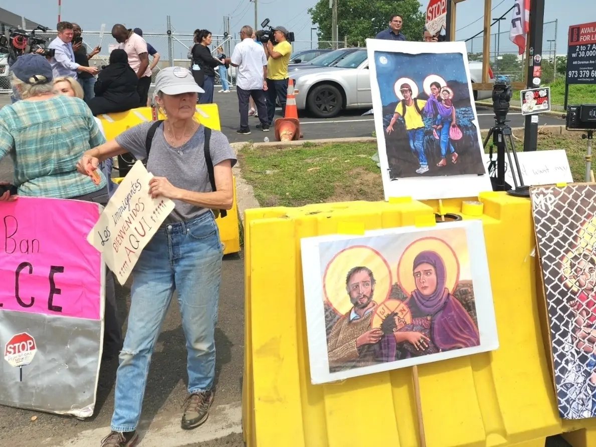 At a vigil outside Delaney Hall, a woman holds a sign reading Los inmigrantes bienvenidos aqui beside paintings of the Holy Family as migrants.