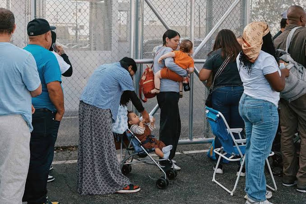 Families wait in line at the tall fence gate, one woman holding a toddler and another bending over a stroller.