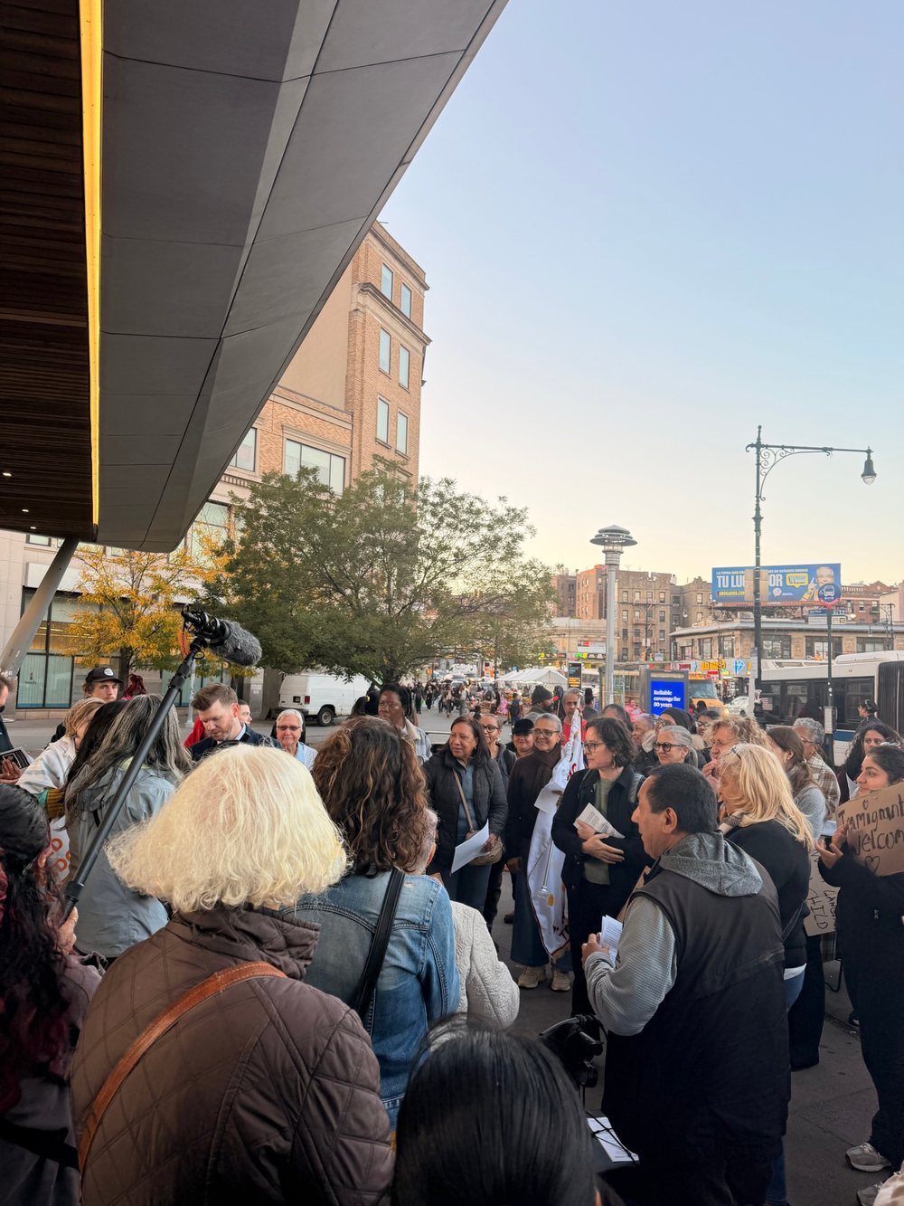 A crowd gathers on a Bronx plaza at sunset before the procession. People hold papers and handmade signs while a man at the front speaks.