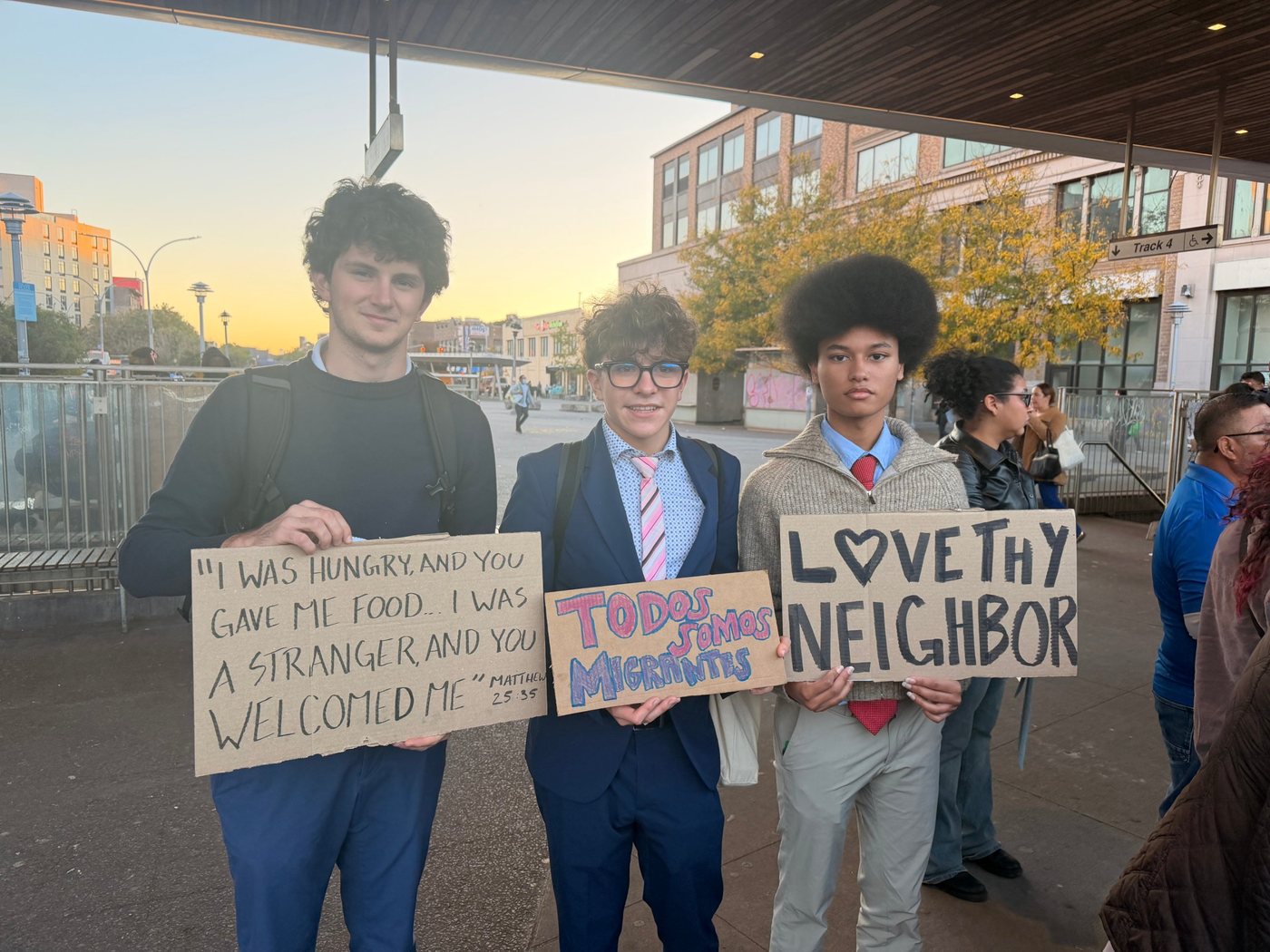Three students in school clothes hold cardboard signs at sunset. One sign quotes Matthew 25:35, another reads Todos somos migrantes and the third reads Love thy neighbor.