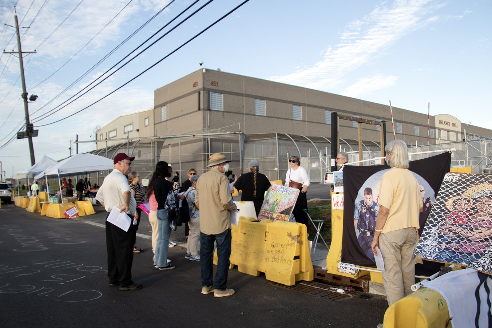 People gather by yellow barriers on the road outside Delaney Hall, a long tan building with its name and the number 451 on the front.